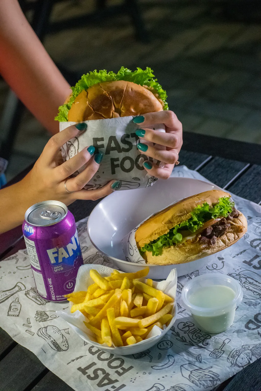 Pessoa segurando um lanche do Mastercheff junto de outro lanche e batata frita